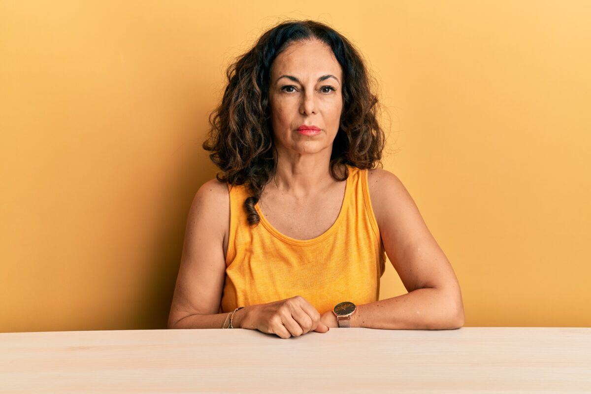 Beautiful middle age woman wearing casual clothes sitting on the table relaxed with serious expression on face. simple and natural looking at the camera.